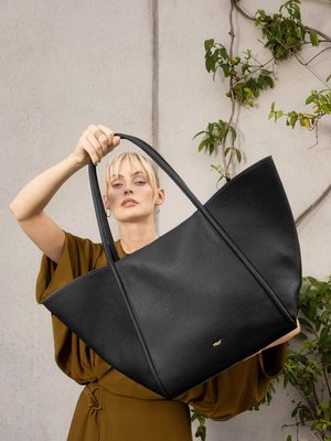 Femme blonde en robe marron tenant un grand sac fourre-tout en cuir noir avec de longues anses, posée contre un mur en béton couvert de vignes vertes.
