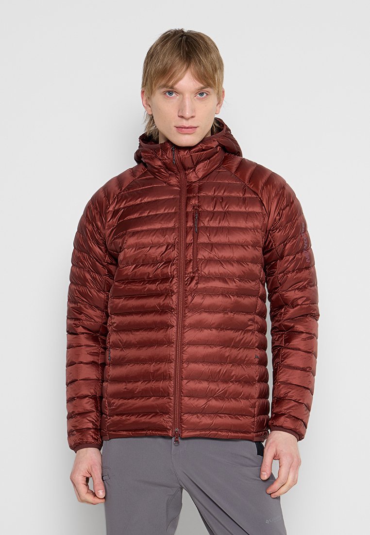 Joven con chaqueta acolchada con capucha de color granate y pantalones grises, de pie frente a un fondo liso de color gris claro.