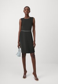 Vestido preto sem mangas com um design ajustado, detalhe branco na cintura, textura suave, combinado com sapatos de salto transparente e uma mala azul clara.