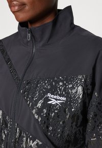 Reebok jacket featuring a high collar, black fabric with textured accents, and a white logo. The design includes diagonal patterns and a zipper.