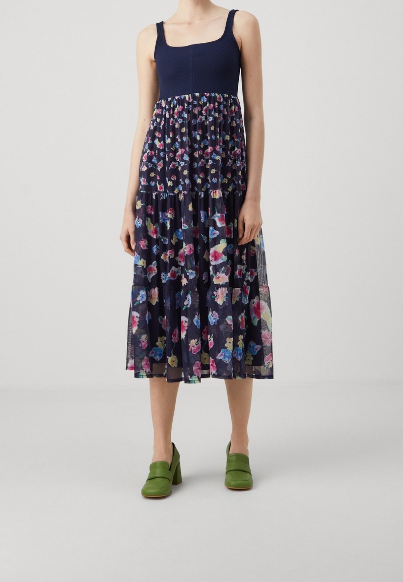Femme portant une robe bleu marine sans manches avec une jupe à motif floral et des chaussures à talons vertes, debout devant un fond uni.