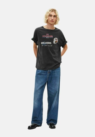 Juodas grafikos marškinėliai su tekstu ir erelio dizainu, derinami su plačiais mėlynais džinsais ir juodais batais. Kasdienis siluetas ir paprastos tekstūros.