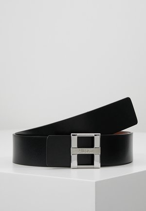 Czarny skórzany pasek z prostokątną srebrną klamrą z wygrawerowanym napisem "HUGO", ułożony w zwiniętą spiralę na białej powierzchni na jednolitym tle.