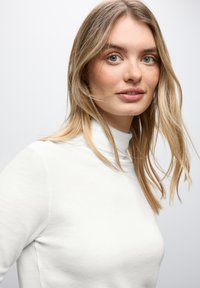 Witte, aansluitende coltrui met een zachte textuur. Heeft een hoge kraag en lange mouwen, gedragen door een model met lang blond haar.