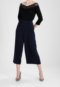 Svart av-axeln-top med spetsdetaljer, matchad med marinblå culottbyxor. Outfiten har strukturerade veck och blanka silvriga högklackade skor.