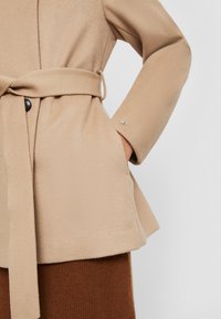 Beige mantel met ceintuur en een gestructureerde afwerking, voorzien van een knoopsluiting en zijzakken. Verfraaid met een klein logo op de manchetten.