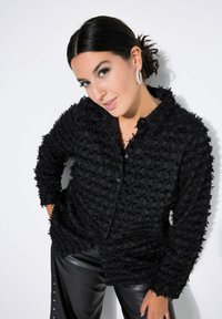 Camicia nera con bottoni in tessuto testurizzato, caratterizzata da dettagli arricciati corti, maniche lunghe e una vestibilità leggermente oversize. Abbinata a pantaloni neri lucidi.