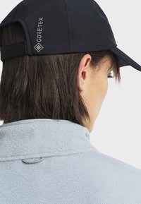 Crna Gore-Tex kapa s podesivom trakom, nosi se uz svijetloplavi flis jaknu s visokim ovratnikom i teksturiranom tkaninom.