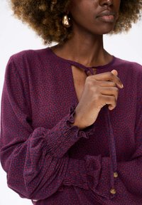 Paarse blouse met kleine rode bloemmotieven, gerimpelde manchetten en smockdetails. Inclusief een gestrikt decolleté en decoratieve kralenaccenten.