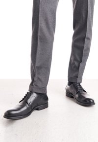 Chaussures habillées en cuir noir avec une finition lisse, bout rond et design à lacets. Portées avec un pantalon gris ajusté avec ourlets.