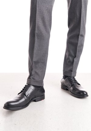 Schwarze Lederschuhe mit glatter Oberfläche, runder Zehenform und Schnürdesign. Getragen mit grauen maßgeschneiderten Hosen mit umgekrempelten Säumen.