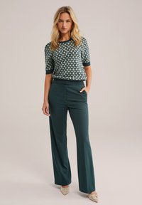 Un pantalon large vert-bleu associé à un pull à manches courtes à motifs vert foncé avec un ourlet côtelé. Les chaussures sont de couleur beige clair avec un détail de boucle.