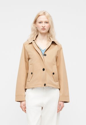 Lyshåret kvinde iført en beige jakke med sorte knapper over en blå top og hvide bukser, stående mod en ensartet lys baggrund.