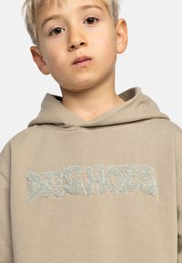 Lichtbeige hoodie met een groot, getextureerd logo in vervaagd groen. Zachte stof, losse pasvorm en een trekkoordkap zorgen voor comfort.