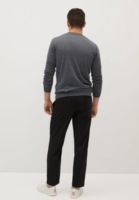 Mann trägt einen grauen Pullover mit langen Ärmeln, schwarze Hosen und weiße Turnschuhe und steht vor einer schlichten weißen Wand.