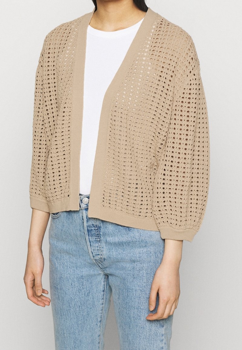 Cárdigan beige de punto con patrón de punto abierto, bordes acanalados y mangas tres cuartos, llevado sobre una camiseta blanca y jeans azul claro.