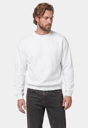 Mand med lysebrunt hår og skæg iført en ensfarvet hvid sweatshirt og mørke jeans, der kigger til venstre mod en ensfarvet grå baggrund.