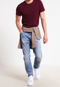 Camiseta burdeos de manga corta, suéter beige atado a la cintura, jeans de mezclilla azul claro y zapatillas blancas. Atuendo casual de corte relajado.