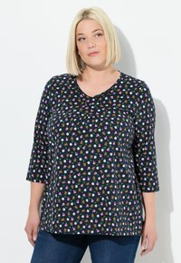 Frau mit blonden Bob-Haar trägt ein schwarzes Langarmoberteil mit kleinen Tier- und Blumenmotiven, kombiniert mit blauen Jeans, und steht vor einer weißen Wand.