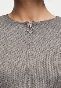 Εγγύς πλάνο ενός γκρι υφασμάτινου ρούχου με ασημένιο κυκλικό φερμουάρ στο λαιμό, που φοριέται από άτομο με ορατό λαιμό και κλείδα.