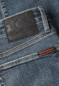 Jeans de mezclilla azul con un parche de cuero negro con diseño en relieve, costuras naranjas y una pequeña etiqueta roja "SUPERDRY" en el bolsillo.