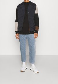 Svart ärmlös puffväst över en stickad tröja med flerfärgade ränder, ihopparad med ljusblå denimjeans och vita sneakers.