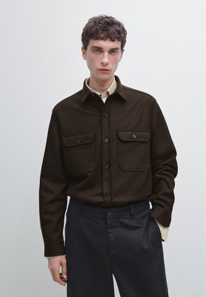 Bruine button-up shirt gemaakt van gestructureerde stof met twee borstzakken. Draagt over een lichtgekleurde hemd met kraag en gecombineerd met donkere broeken.