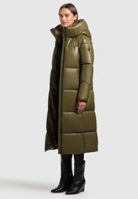 Olijfgroen pufferjack met een hoge kraag, een geborduurd patroon en een ritsluiting. Het model draagt zwarte kniehoge haklaarzen.