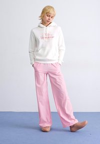 Witte hoodie met roze logo, gecombineerd met losse roze broek en beige clogs. Gladde stof en ontspannen pasvorm ontwerp.