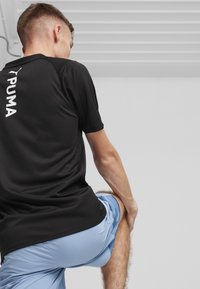 Must neljas must sportlik särk, millel on võrgumuster ja seljal silmatorkav valge "PUMA" logo, sobitatud helesiniste lühikeste pükstega.