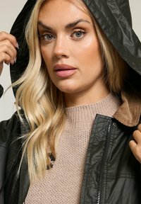 Veste à capuche noire avec une texture matelassée, dotée d'un col en velours côtelé marron, portée sur un pull en maille beige clair.