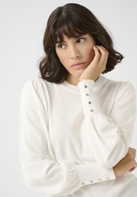 Bílý pletený svetr s nabíranými dlouhými rukávy a přiléhavými manžetami, které mají dekorativní stříbrné knoflíky. Lehká tkanina s hladkou strukturou.