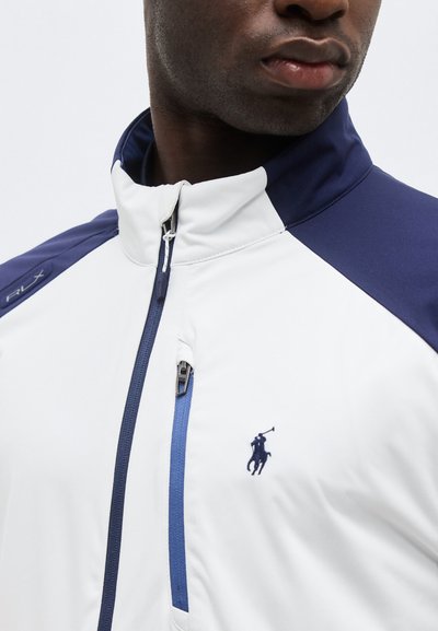 Hombre con chaqueta blanca y azul marino con cremallera, con un logo de jugador de polo en el pecho y un bolsillo con cremallera en el pecho.