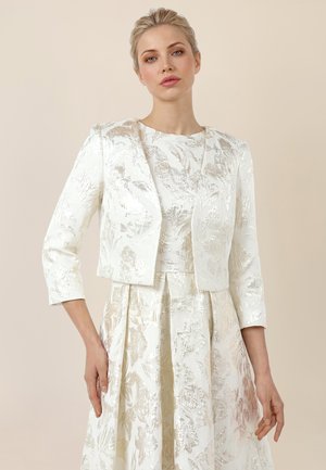 Frau trägt ein weißes Jacquardkleid mit Blumenmuster und eine passende cropped Jacke, steht vor einem neutralen beigefarbenen Hintergrund.