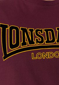 Burgundy pamut póló, nagy fekete és sárga "LONSDALE LONDON" felirattal. Puha tapintású, kerek nyakú kialakítással.