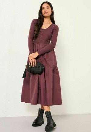 Frau in einem langärmeligen, mauvefarbenen Kleid, die eine kleine schwarze Tasche hält und mit schwarzen Stiefeletten kombiniert ist, steht vor einer schlichten, elfenbeinfarbenen Wand.