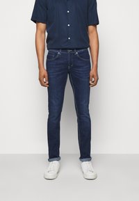 Vaqueros de mezclilla azul oscuro con un corte ajustado, con costuras en contraste, cinco bolsillos y dobladillo enrollado, combinados con zapatillas blancas.