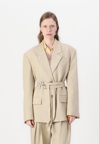 Beżowy oversize'owy żakiet z szerokimi ramionami, w talii z paskiem i bocznymi kieszeniami. Nosi się go na jasnożółtą koszulę o lekkiej teksturze i strukturalnym designie.