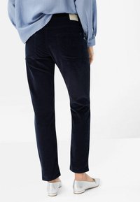 Personne portant un pantalon bleu marine foncé et un haut bleu clair, vue de dos, avec des chaussures plates argentées sur fond blanc.