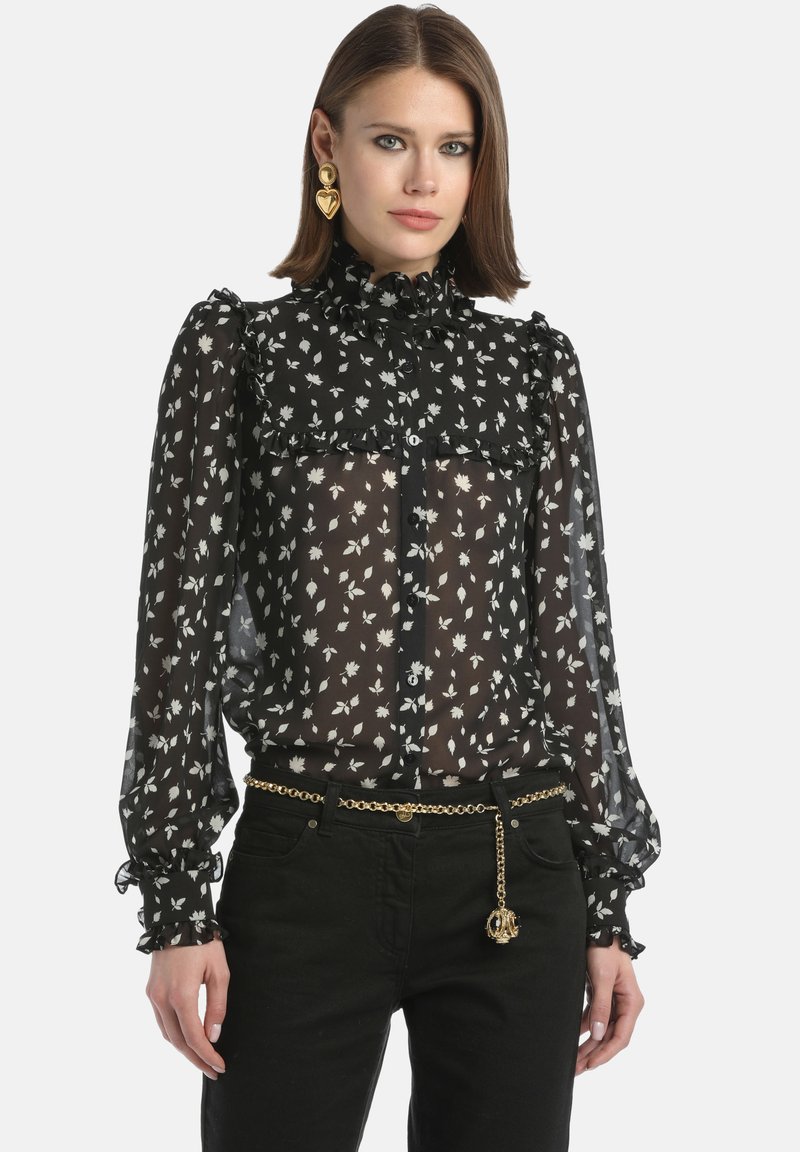 Black sheer blouse with white floral print, ruffled collar, and button front. Long sleeves with ruffle cuffs. Paired with black pants and gold chain belt.