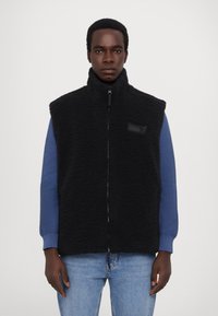 Black fleece vest with high collar, front zipper, and logo patch, paired with a blue knit long-sleeve shirt. Denim jeans complete the look.