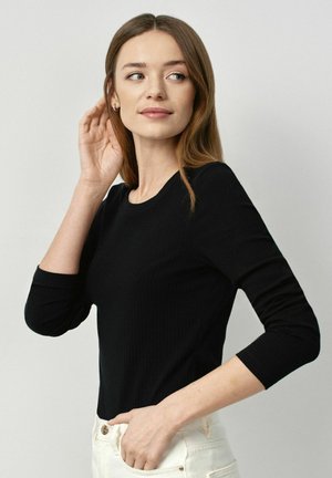Czarna prążkowana bluzka z długim rękawem i okrągłym dekoltem. Materiał wydaje się miękki, o dopasowanym kroju, noszona z jasnymi spodniami.