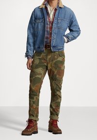 Chaqueta de mezclilla con cuello beige, camisa de cuadros, pantalones cargo de camuflaje con acentos bordados y botas marrones con cordones. Incluye bolsillos y puños enrollados.