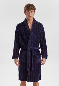 Accappatoio a righe blu navy con linee rosse, realizzato in morbido tessuto di spugna, presenta un colletto a scialle, due tasche frontali e una cintura abbinata.