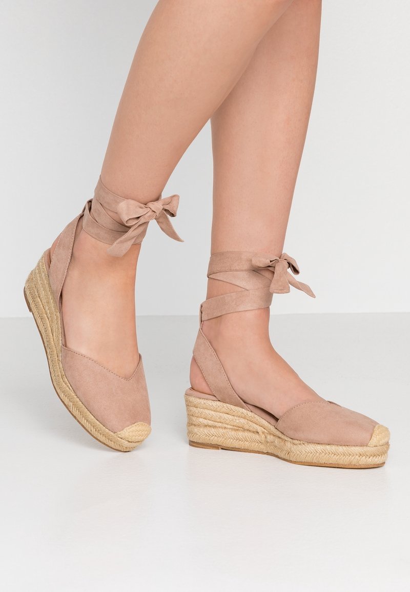 Jambes portant des espadrilles compensées beiges avec des liens à nouer autour de la cheville, debout sur un sol blanc uni contre un fond blanc.