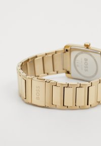 Montre-bracelet en métal doré avec un boîtier rectangulaire et un bracelet à maillons. Présente une finition polie et un logo de marque gravé sur le fermoir.