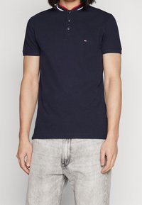 Man klädd i en marinblå kortärmad pikétröja med tre knappar och ljusgrå urtvättade jeans, stående framför en neutral bakgrund.