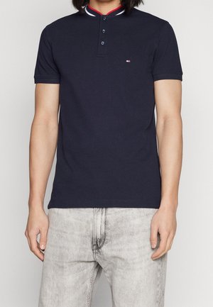 Man in een marineblauwe polo met korte mouwen en drie knopen, met lichtgrijze vervaagde jeans, staande tegen een neutrale achtergrond.