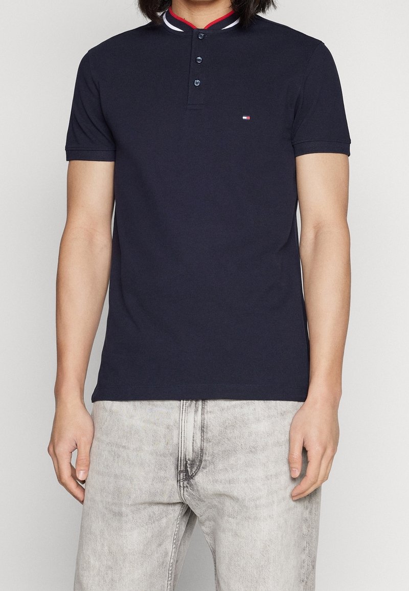 Man klädd i en marinblå kortärmad pikétröja med tre knappar och ljusgrå urtvättade jeans, stående framför en neutral bakgrund.