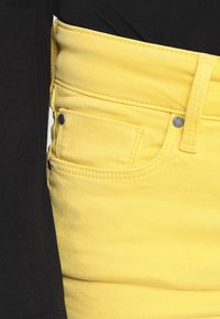 Jeans de denim amarillos con un diseño ajustado. Las características incluyen un bolsillo frontal, botón de metal y una tela ligeramente texturizada. Camiseta negra visible.